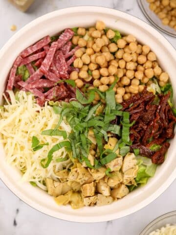 A bowl of chopped salad, featuring mozzarella cheese, artichokes, sun dried tomatoes, chickpeas, salami, and basil.