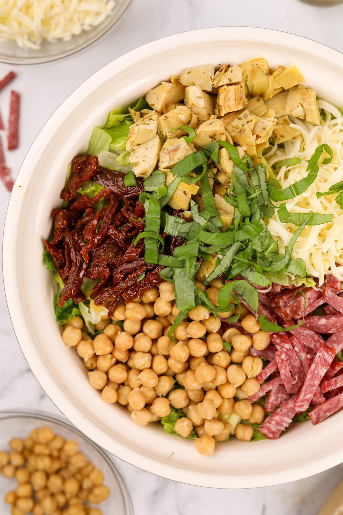 A large bowl of salad, topped with sections of chickpeas, sundried tomatoes, basil, artichokes, mozza, and salami.