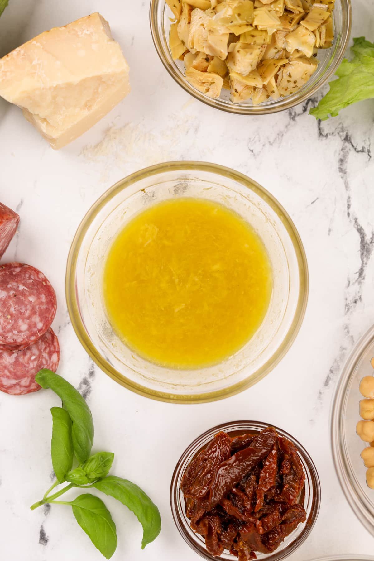 A small bowl of bright vinaigrette, surrounded by antipasto ingredients.