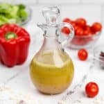 A glass cruet filled with homemade salad dressing, with red pepper, tomatoes, and lettuce in the background.