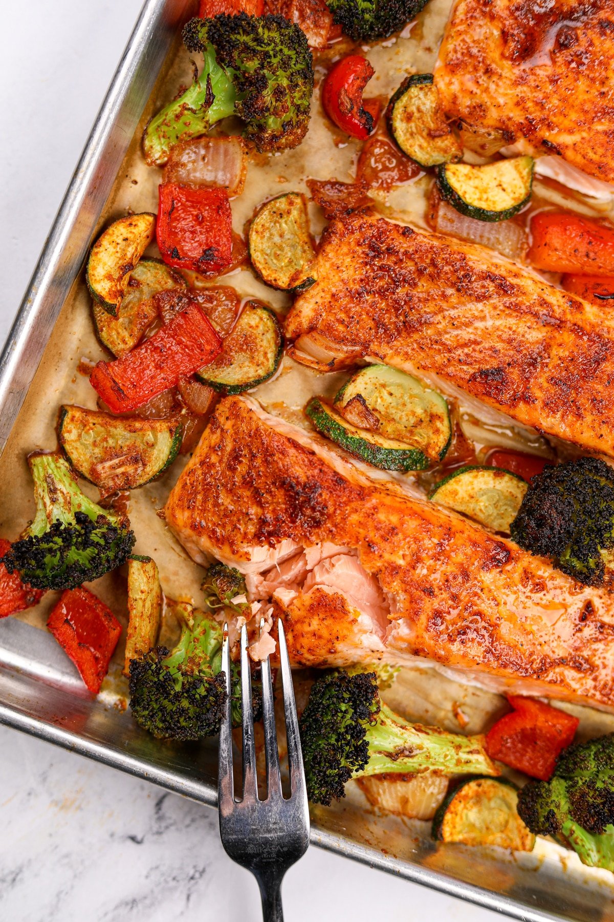 A fork flaking a fillet of salmon, surrounded by various vegetables including broccoli, bell pepper, and zucchini.