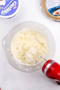 An electric mixer blending cream cheese with sugar.