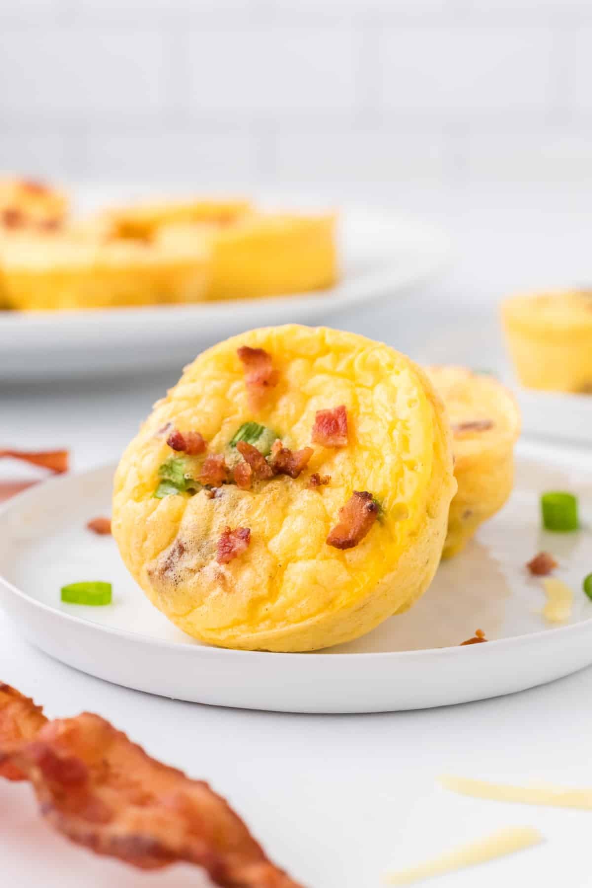Two egg bites on a plate garnished with cooked bacon and green onion.