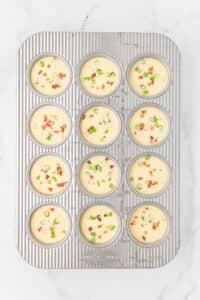 A muffin pan with all of the wells filled with an egg and bacon mixture and sprinkled with green onions.
