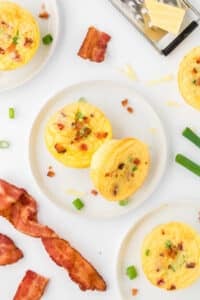 Two egg bites on a white plate, surrounded by more egg bites and strips of bacon.
