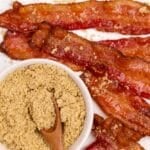 Strips of bacon on a white counter, with a small bowl of brown sugar nearby.