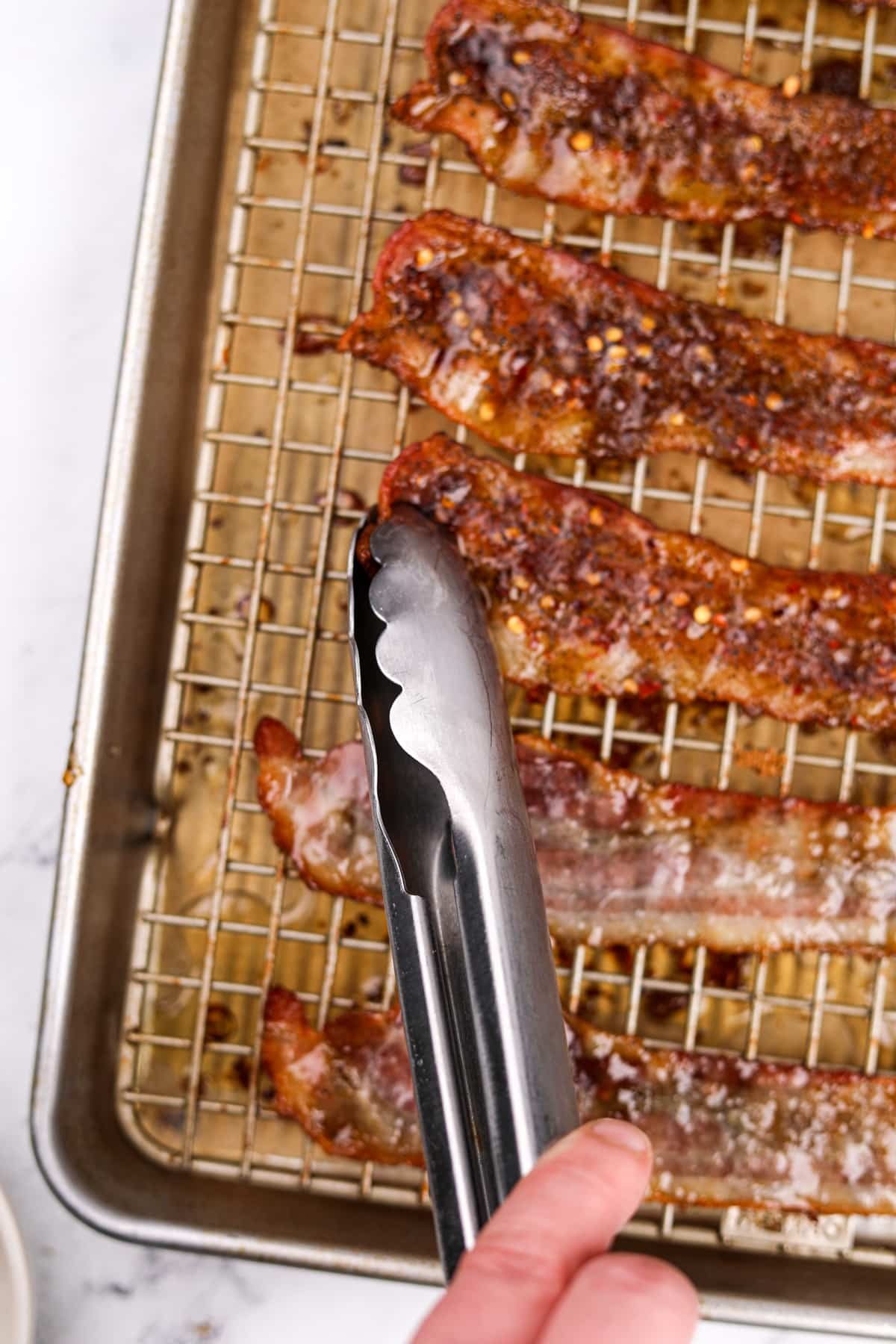 A pair of tongs flipping bacon on a baking sheet.