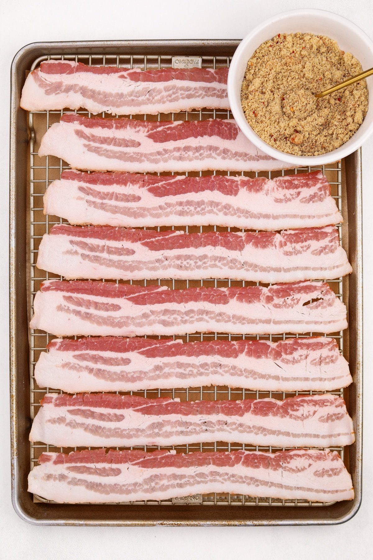 A baking sheet with bacon slices spread out, with a small bowl of brown sugar on the side.