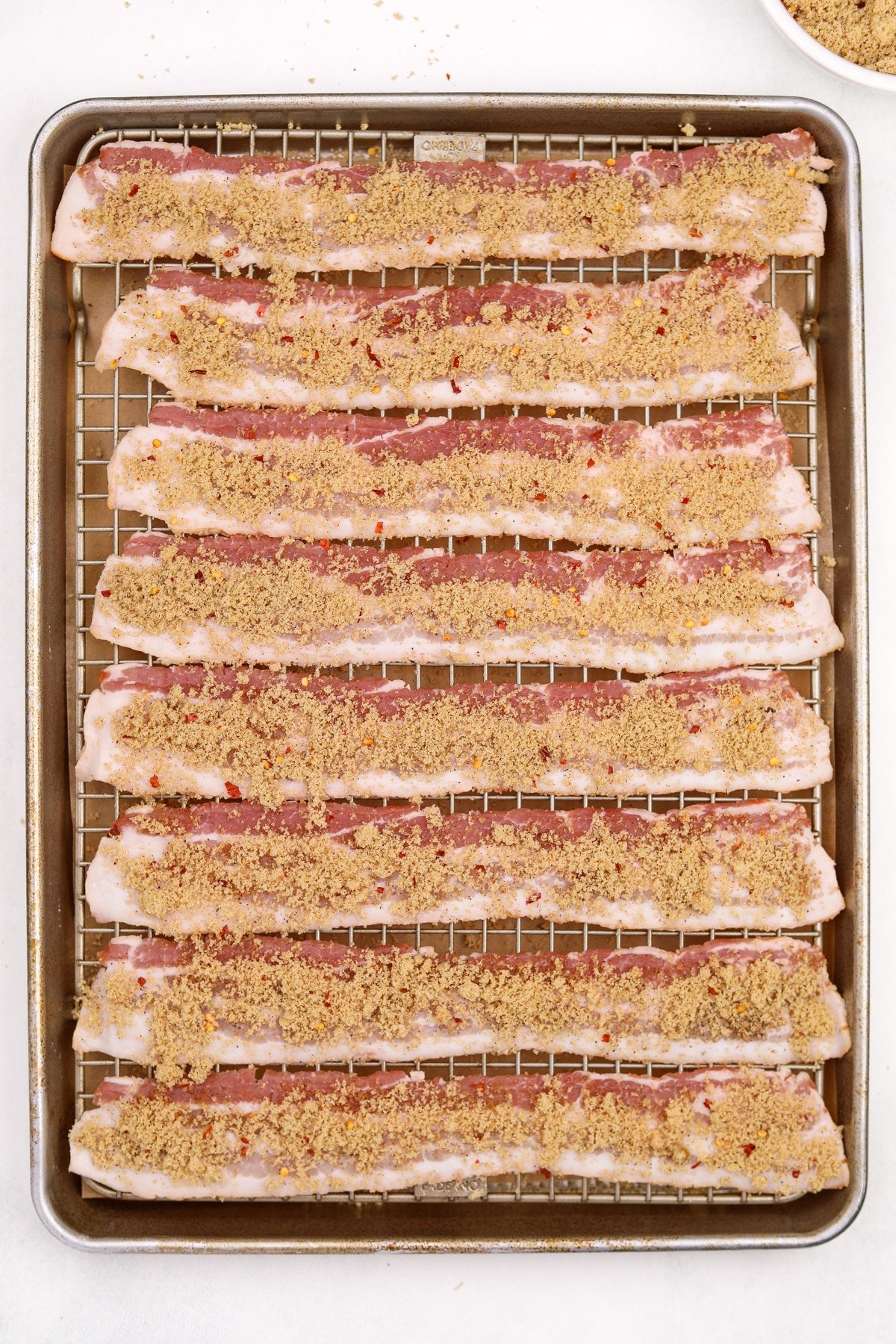 A baking sheet with bacon spread out, topped with a brown sugar rub.
