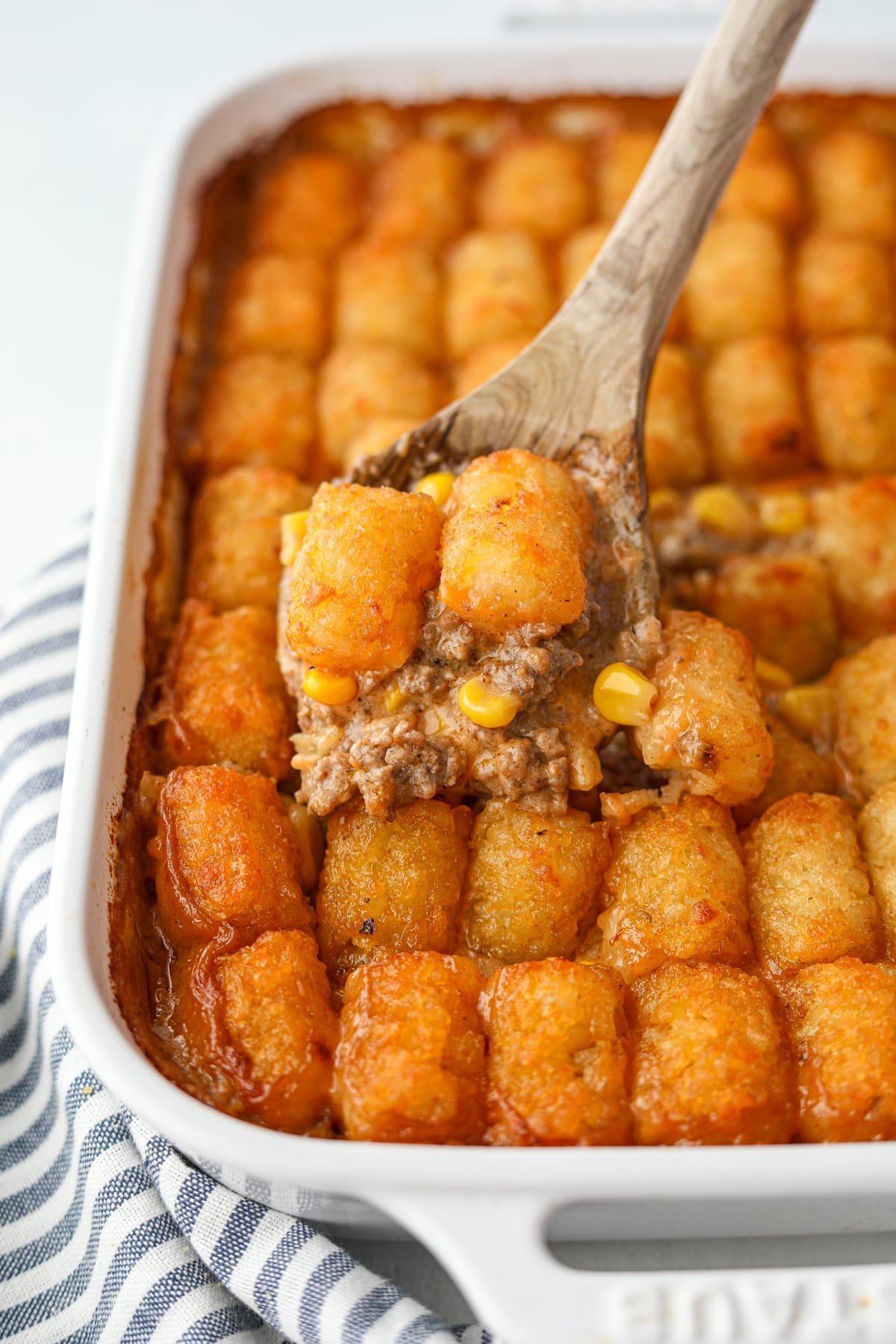 A wooden spoon lifting a Tater Tot topping with some ground beef from a baking dish.