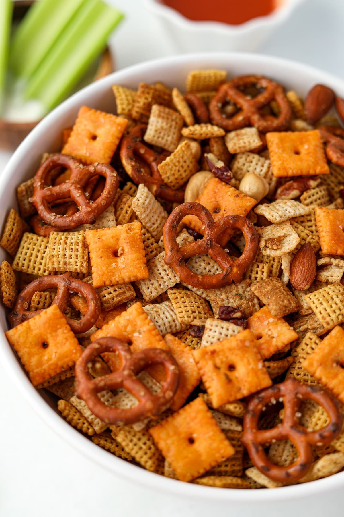 A close up on a bowl of homemade Chex Mix, with celery and buffalo sauce in the background.