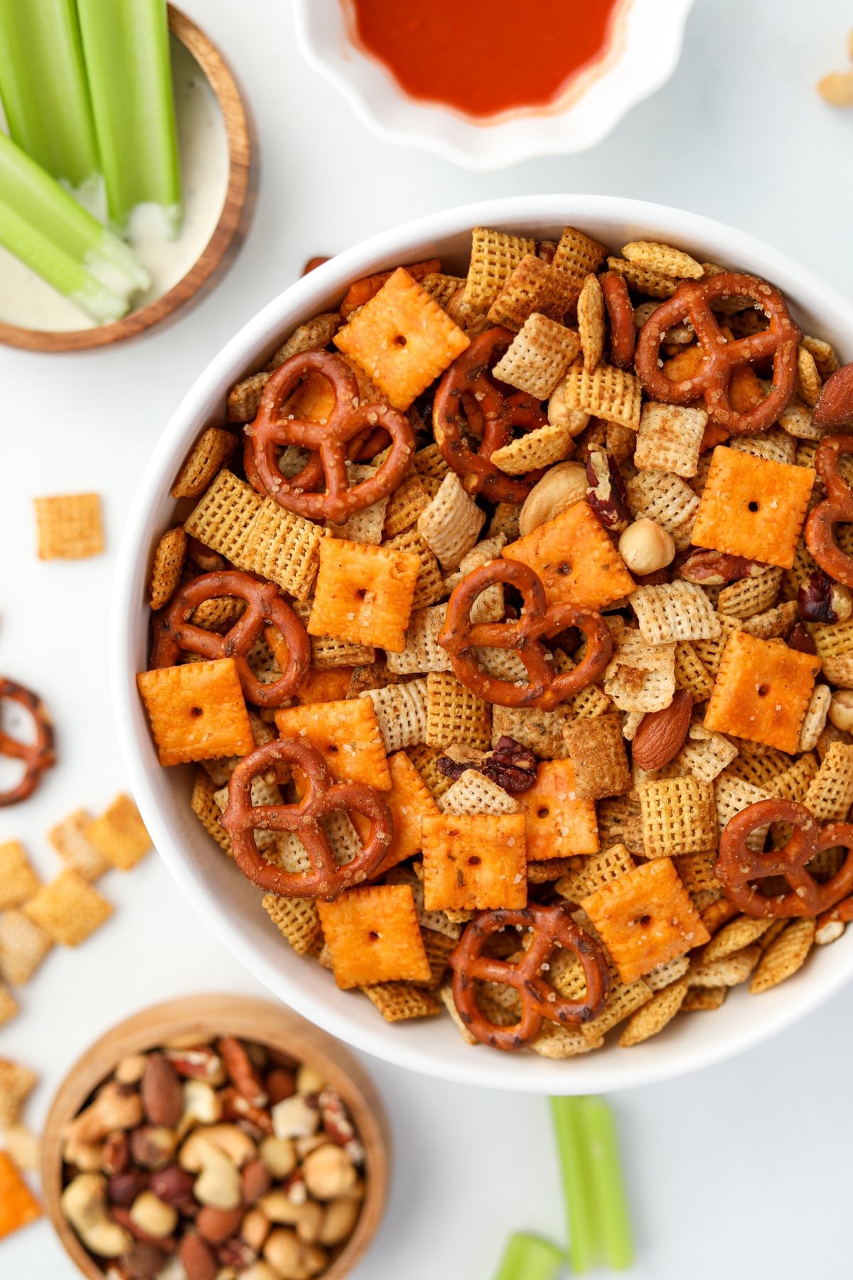 An overhead view of a bowl of Chex mix, with celery, nuts, and buffalo sauce in the background.