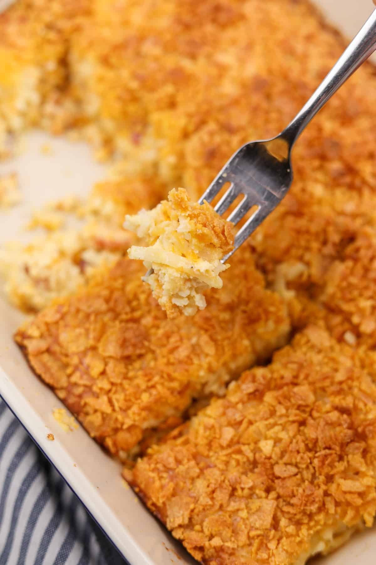 A fork showing the texture of a shredded hashbrown casserole, which is in the background and topped with crunchy cornflakes.