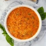 A bowl of cherry tomato soup garnished with parmesan cheese, basil, and cracked black pepper.