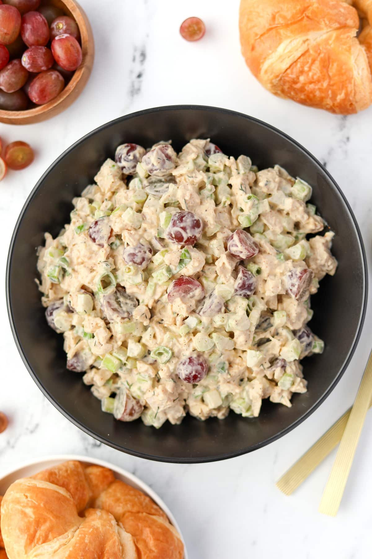 A bowl of chicken salad in a black bowl, surrounded by a bowl of grapes and some croissants. 