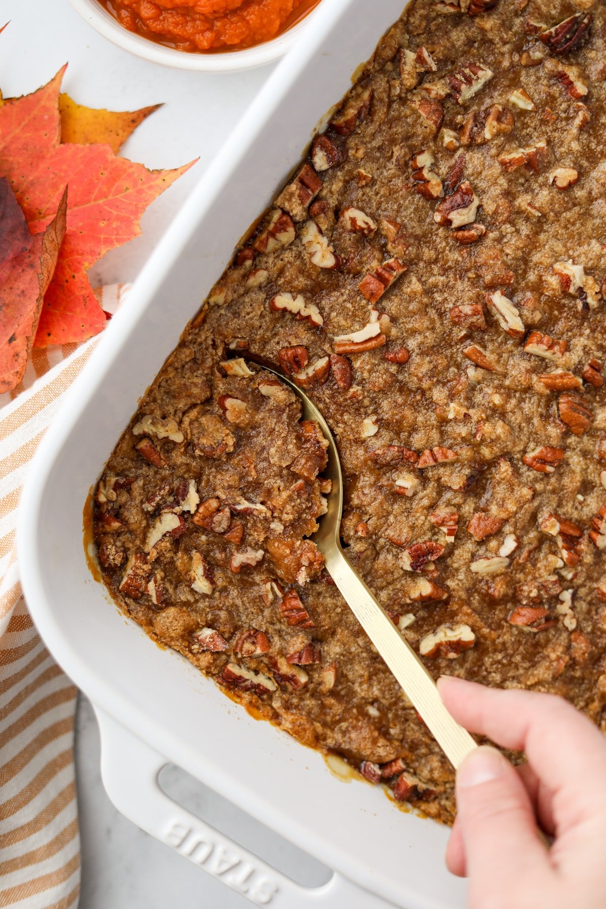 A pecan streusel topped casserole, with a gold serving spoon taking a portion.