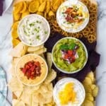 A board featuring bowls of dip and a variety of chips.