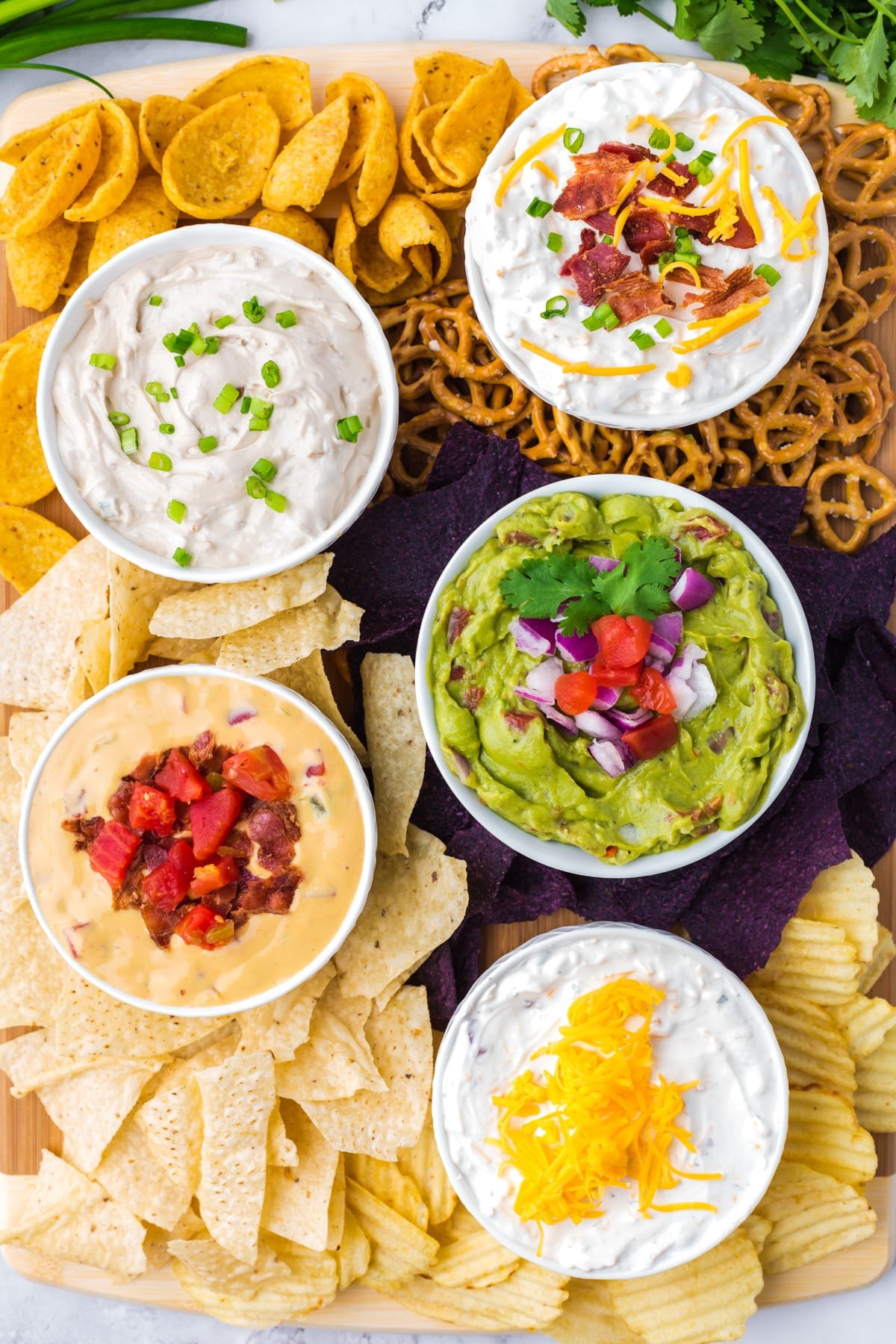 Five small bowls filled with dip, surrounded by various types of chips on a wooden board.