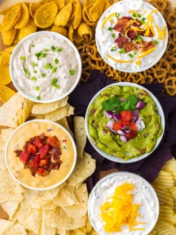 A board featuring bowls of dip and a variety of chips.