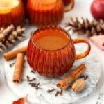 A pumpkin shaped mug filled with homemade cider. Nutmeg, cinnamon sticks, and cloves are on the marble below.