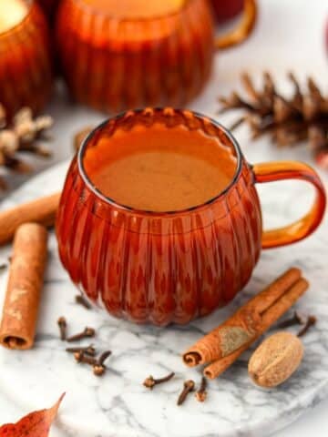 A pumpkin shaped mug filled with homemade cider. Nutmeg, cinnamon sticks, and cloves are on the marble below.