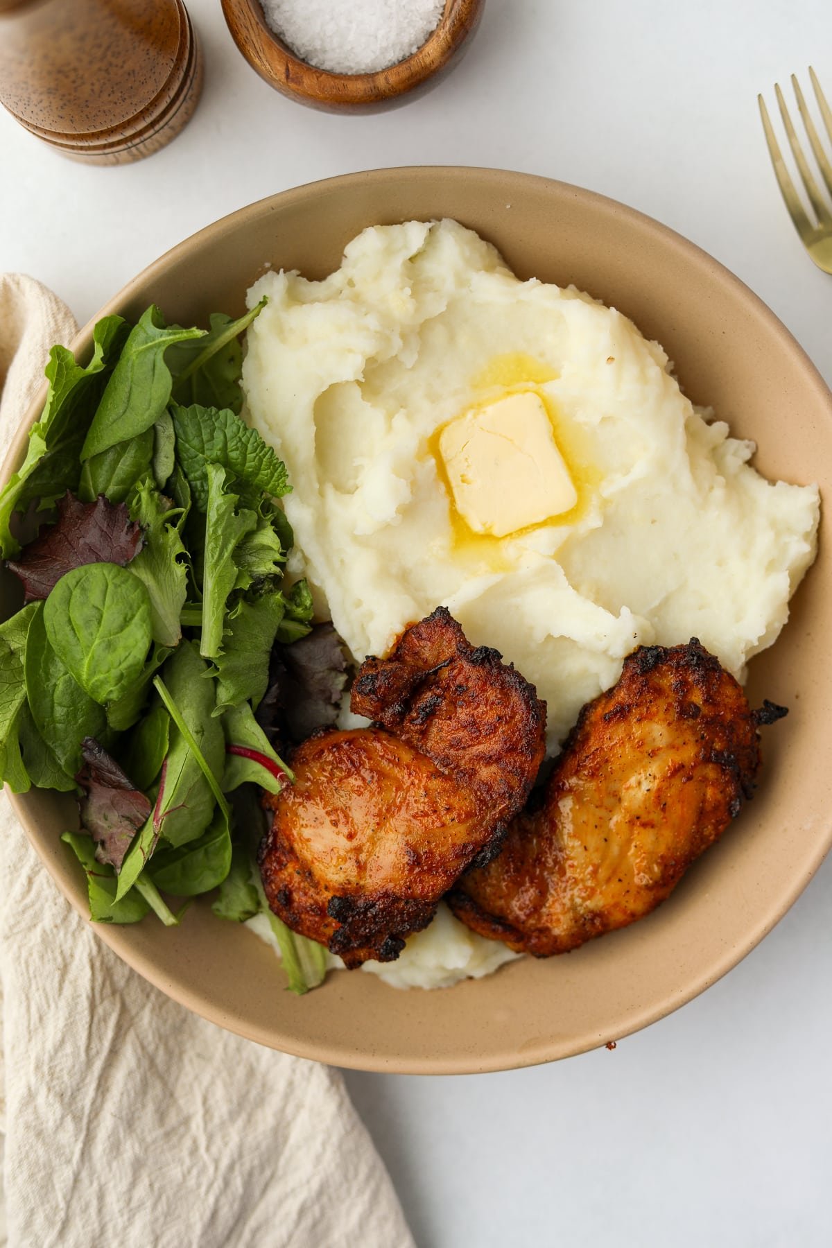 A bowl featuring mashed potatoes, salad, and some crispy chicken thighs.