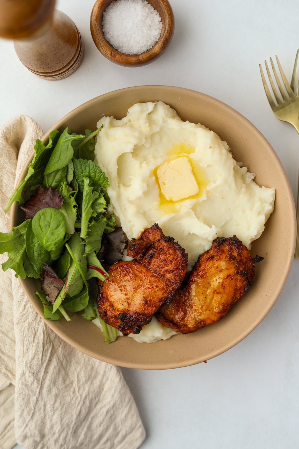 A shallow bowl featuring salad, mashed potatoes, melted butter, and crispy chicken thighs.