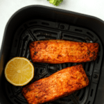 An air fryer basket with two salmon fillets and a round of fresh lemon.