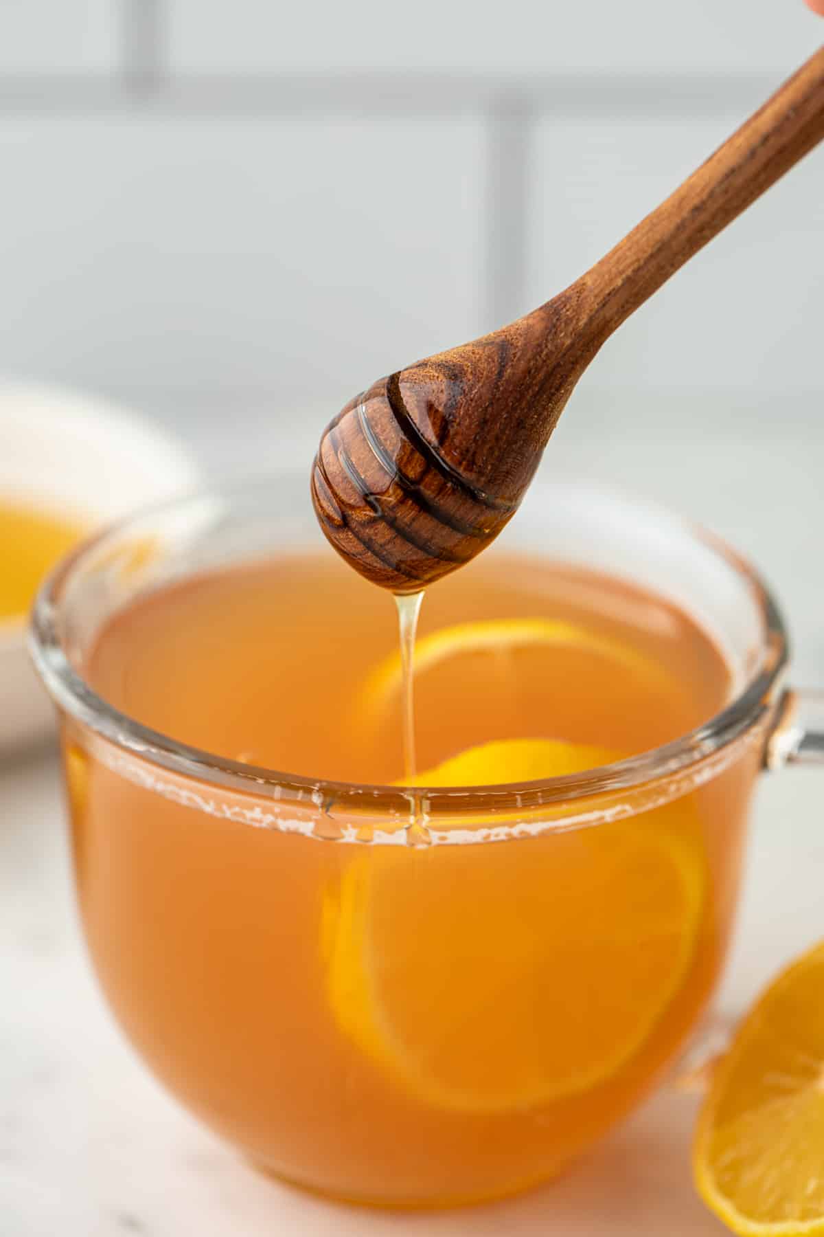 Drizzling honey in a glass of steeped tea garnished with lemon slices.
