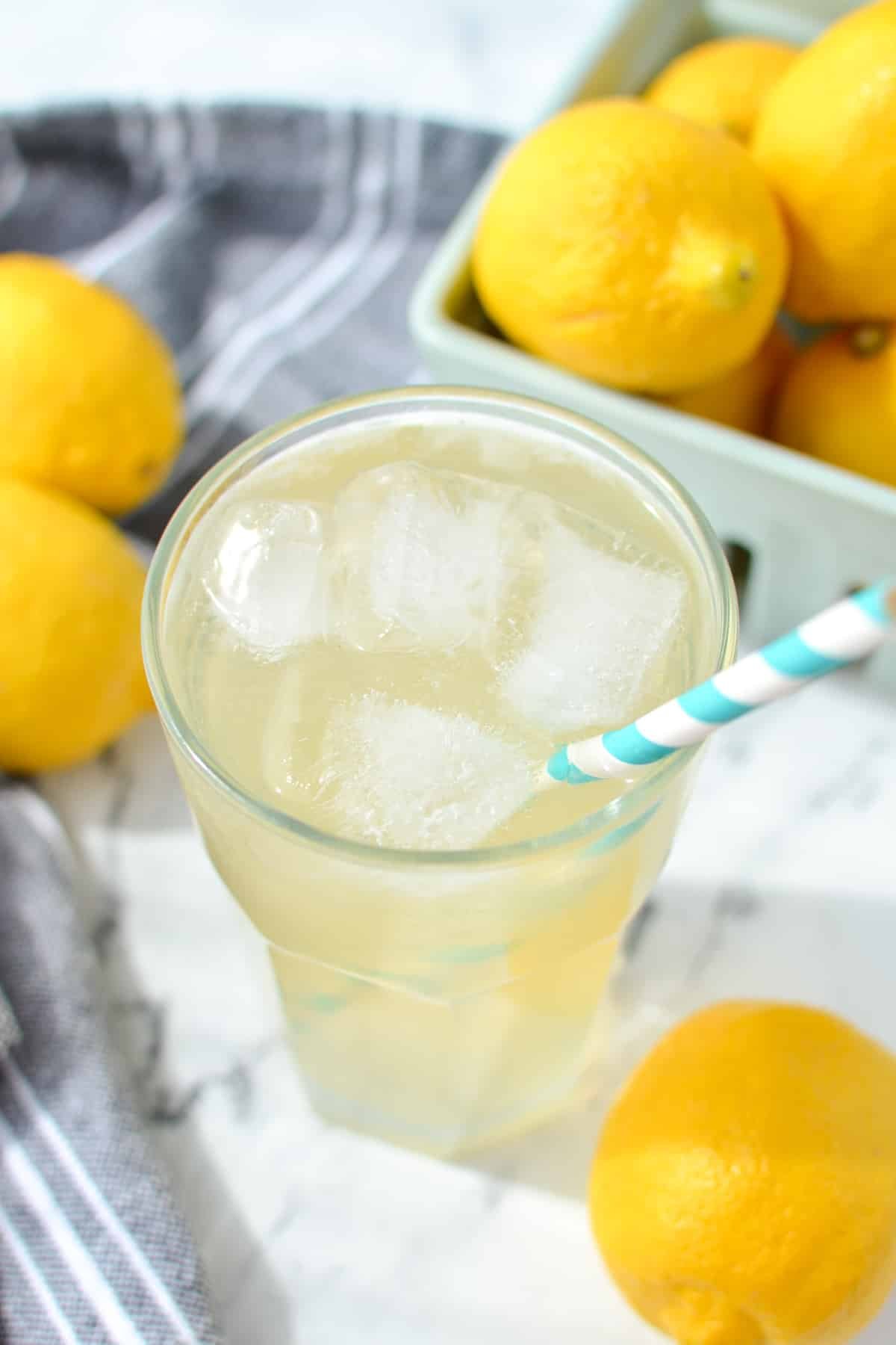 Lemonade in a glass filled with ice, with fresh lemons in the background.