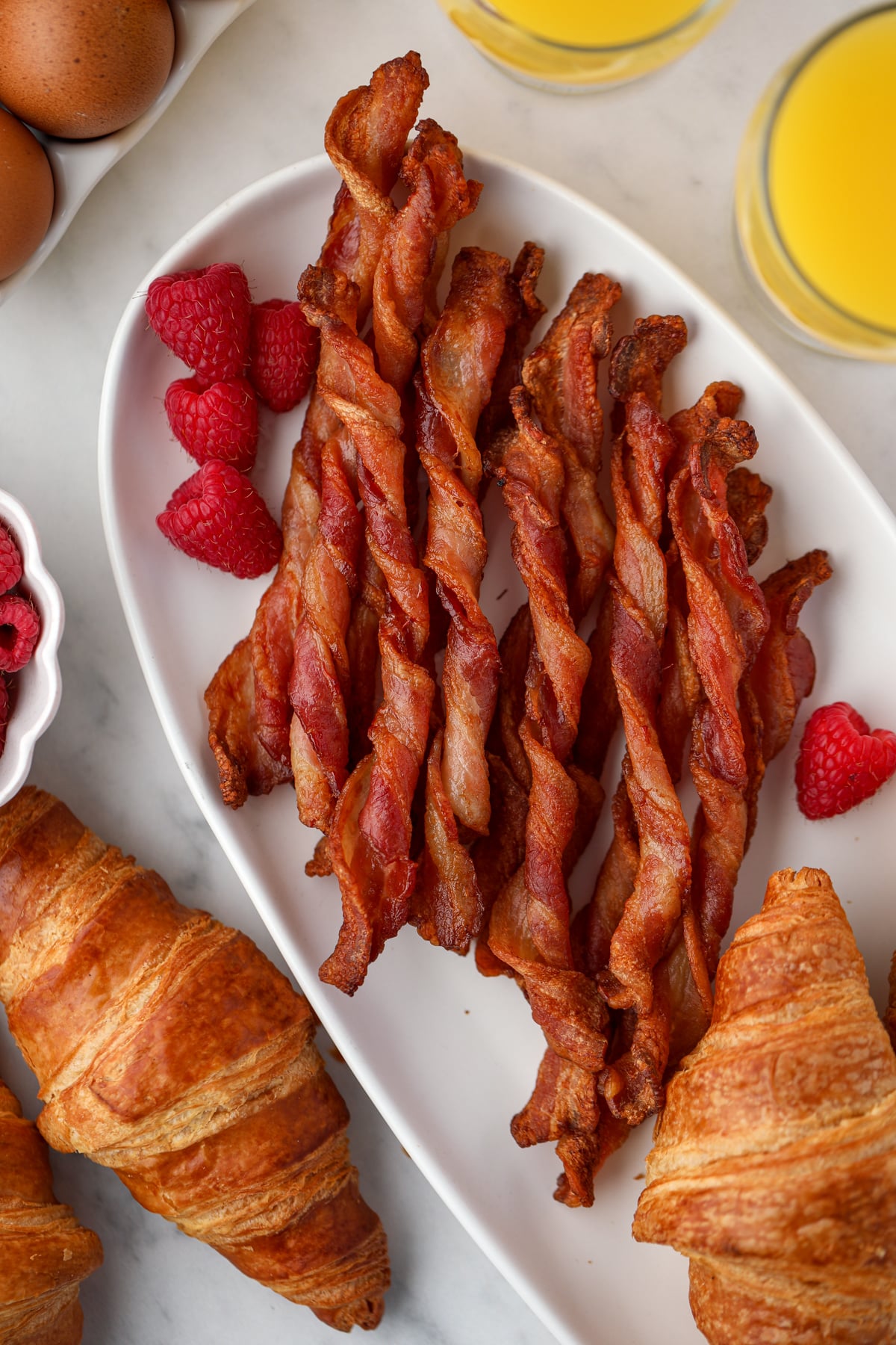 Crispy twisted bacon on a white platter, garnished with fresh raspberries and some croissants on the side.