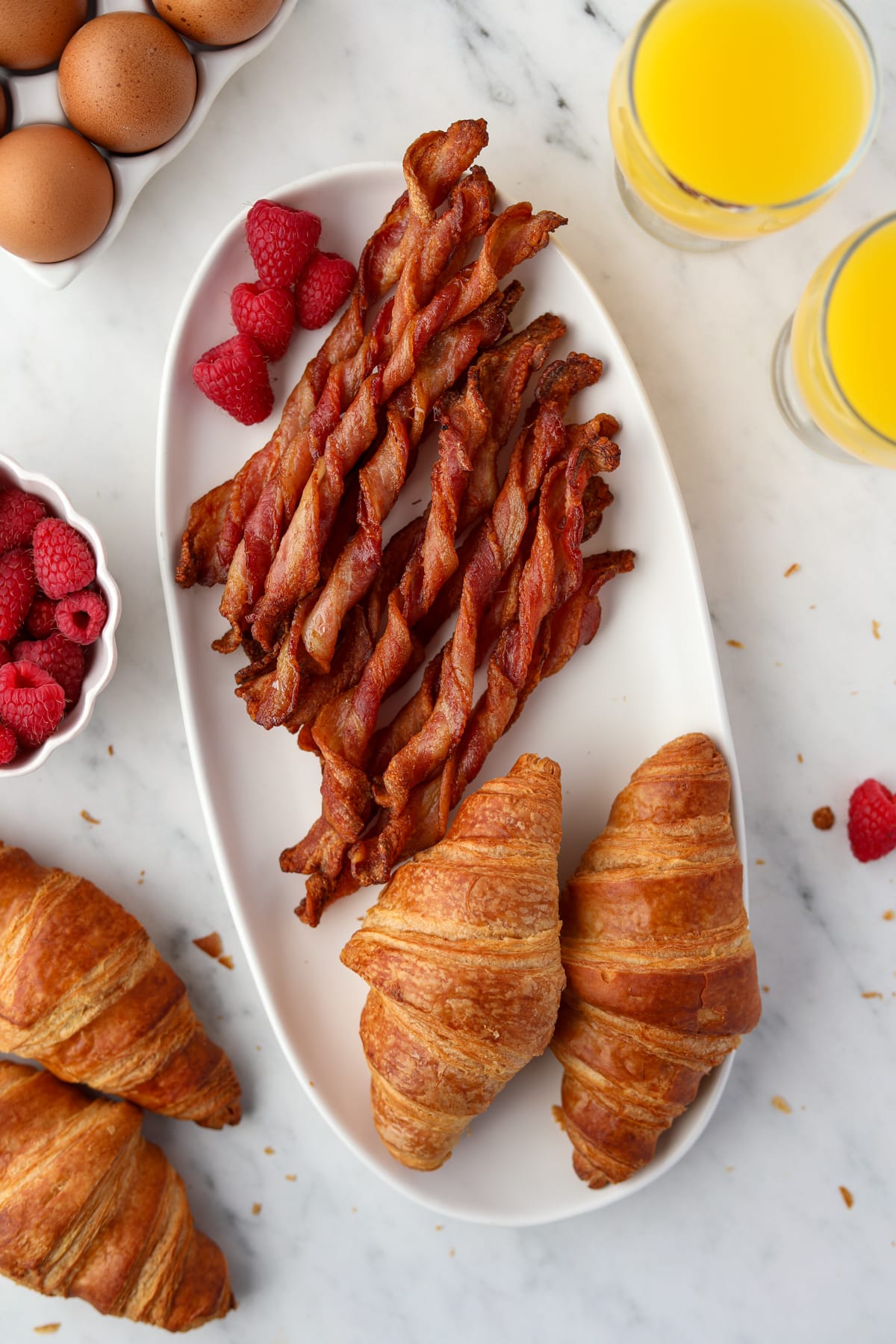 A birds-eye view of a counter featuring a platter of bacon twists and croissants, with the outside edges showing glasses of orange juice and fresh raspberries.