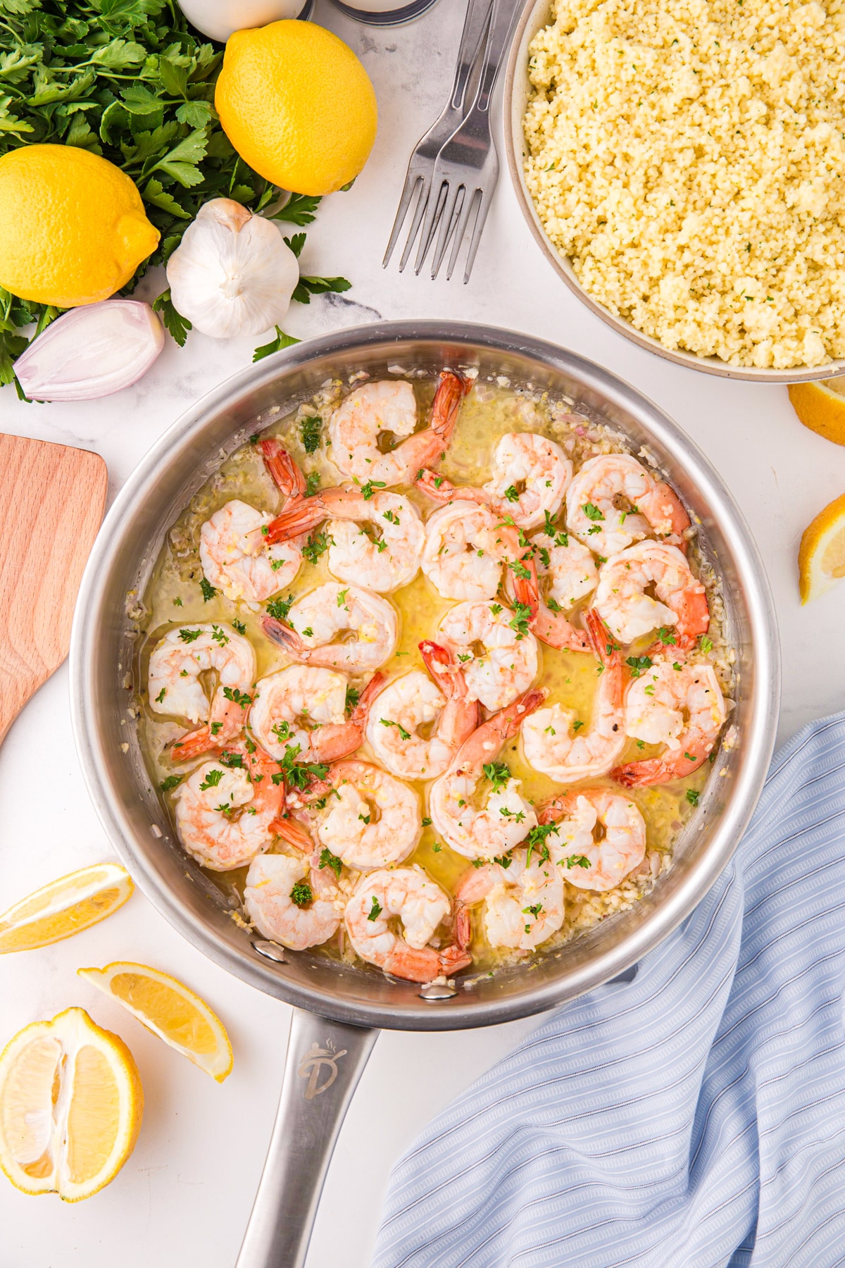 A stainless skillet filled with cooked shrimp; fresh parsley and lemon are on the outsides of the frame.