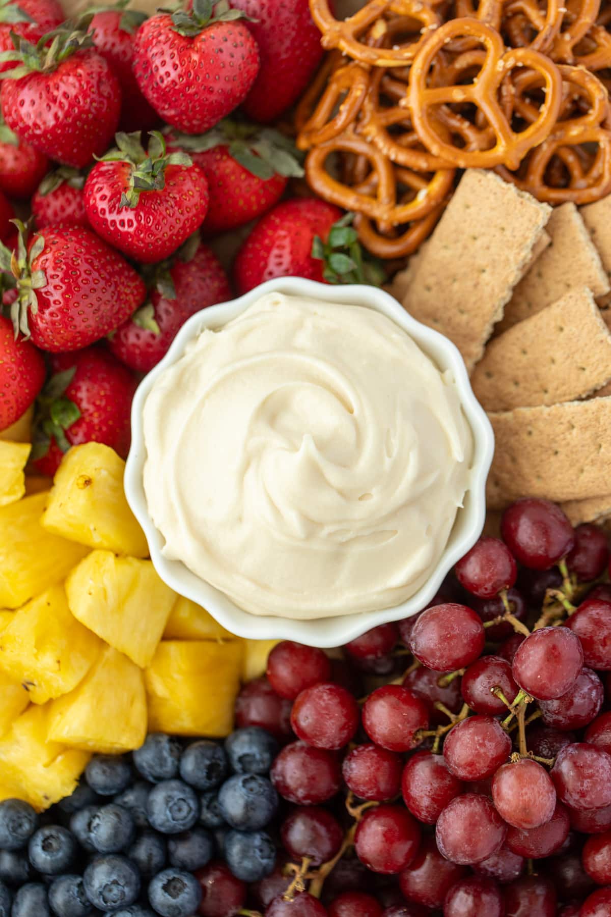 A small bowl of cream cheese dip surrounded by fresh fruit, graham crackers, and pretzels.