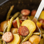 A ladle scooping up potatoes with green beans and sliced sausage.