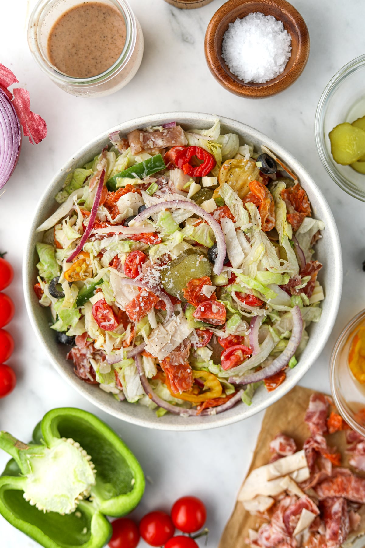 An overhead image of a bowl of salad, surrounded by vegetables and other various toppings.