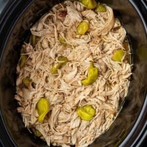 A slow cooker filled with shredded chicken and topped with pepperoncini peppers.