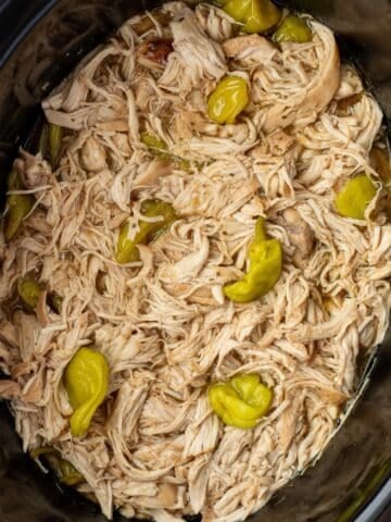 A slow cooker filled with shredded chicken and topped with pepperoncini peppers.