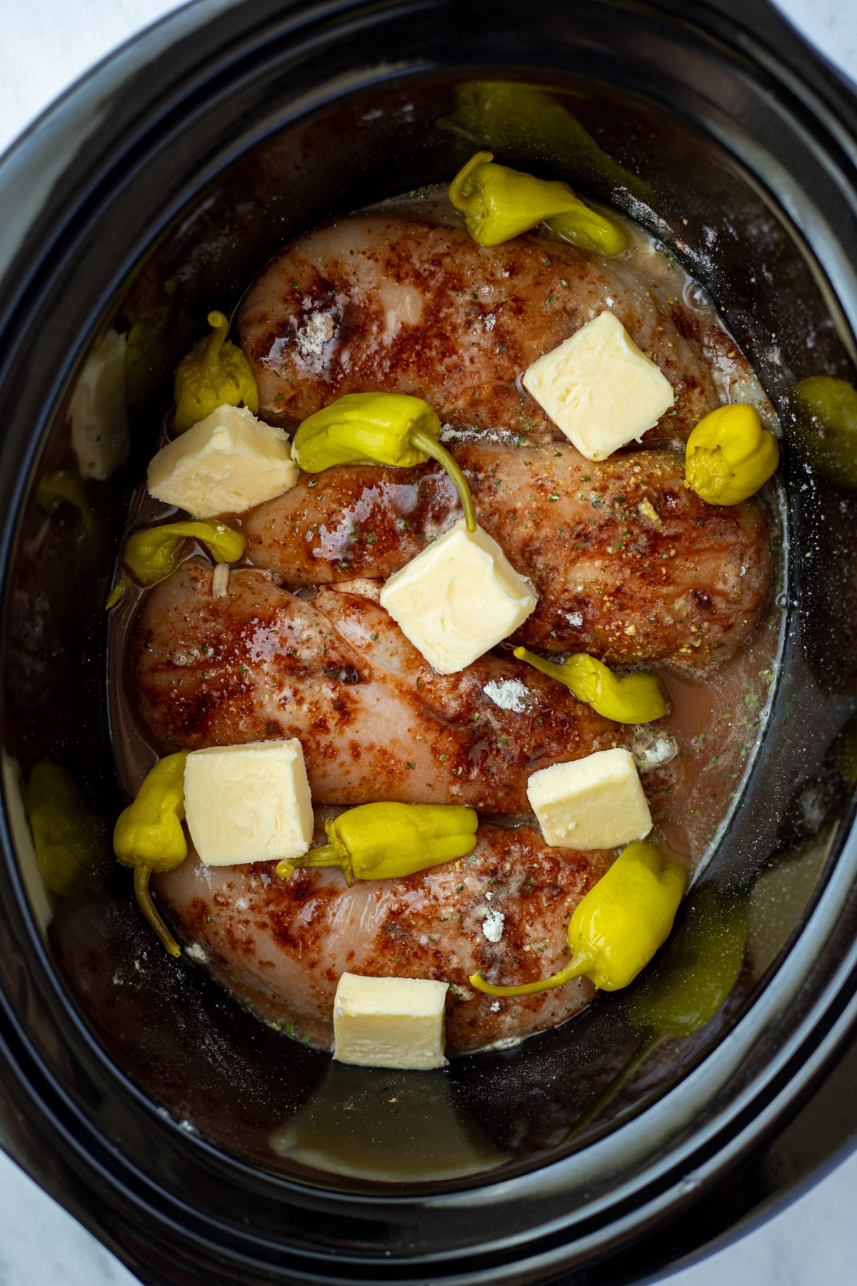 A slow cooker with chicken, seasoning, butter, and pepperoncini peppers.