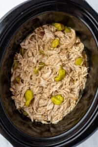 A slow cooker filled with juicy shredded chicken and a few pepperoncini peppers.
