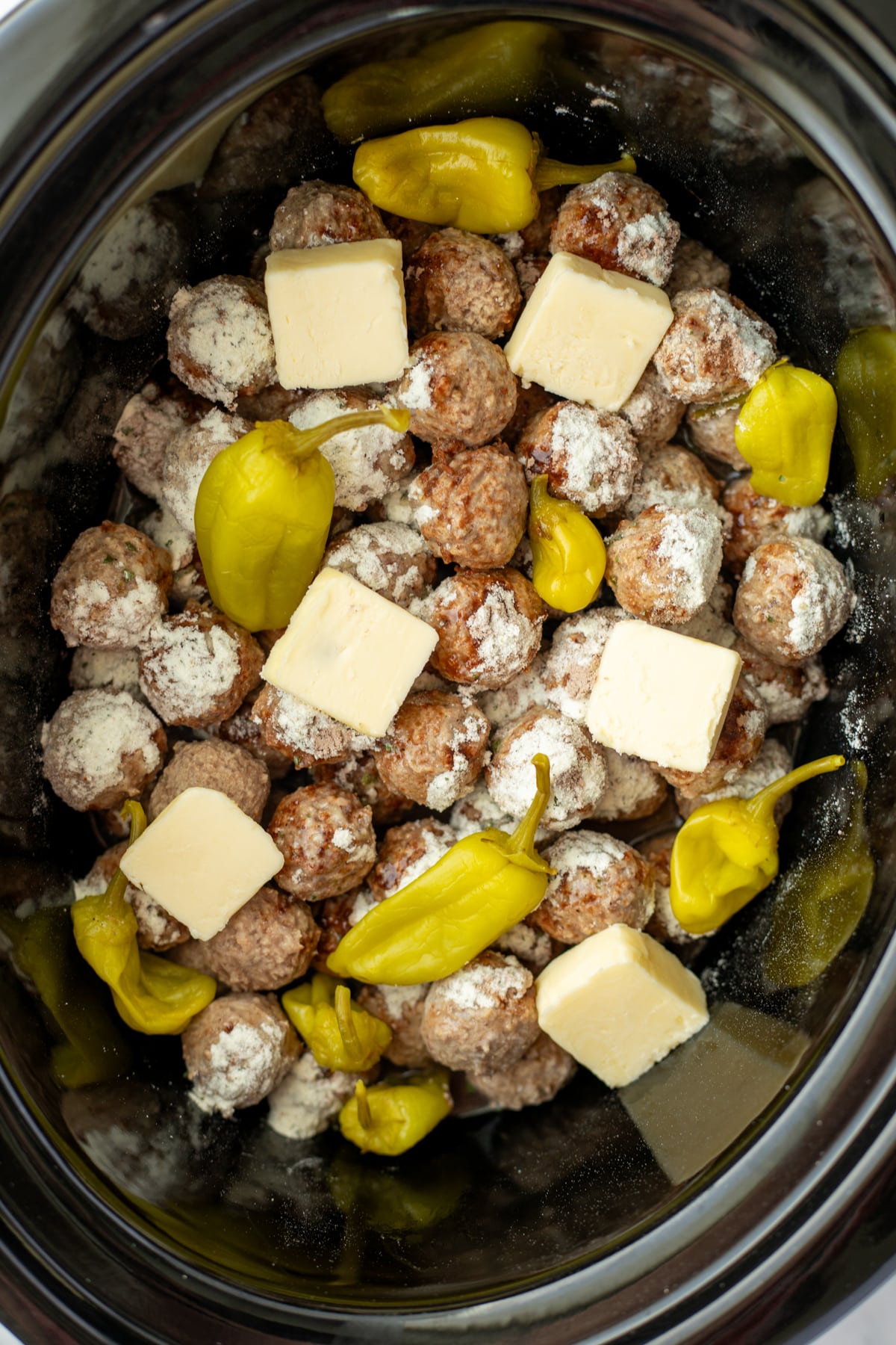 A slow cooker filled with meatballs, pepperoncini peppers, and butter.