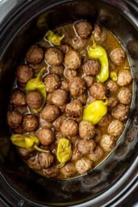 A slow cooker filled with cooked meatballs mixed with a thin sauce and pepperoncini peppers.
