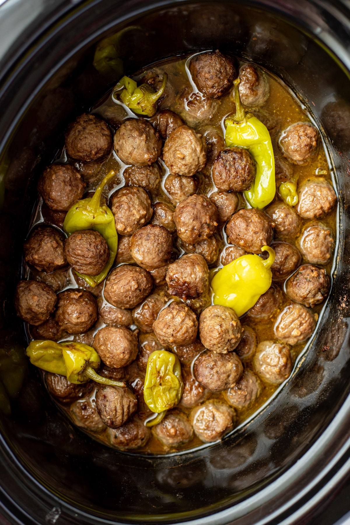 A slow cooker filled with cooked meatballs mixed with a thin sauce and pepperoncini peppers.