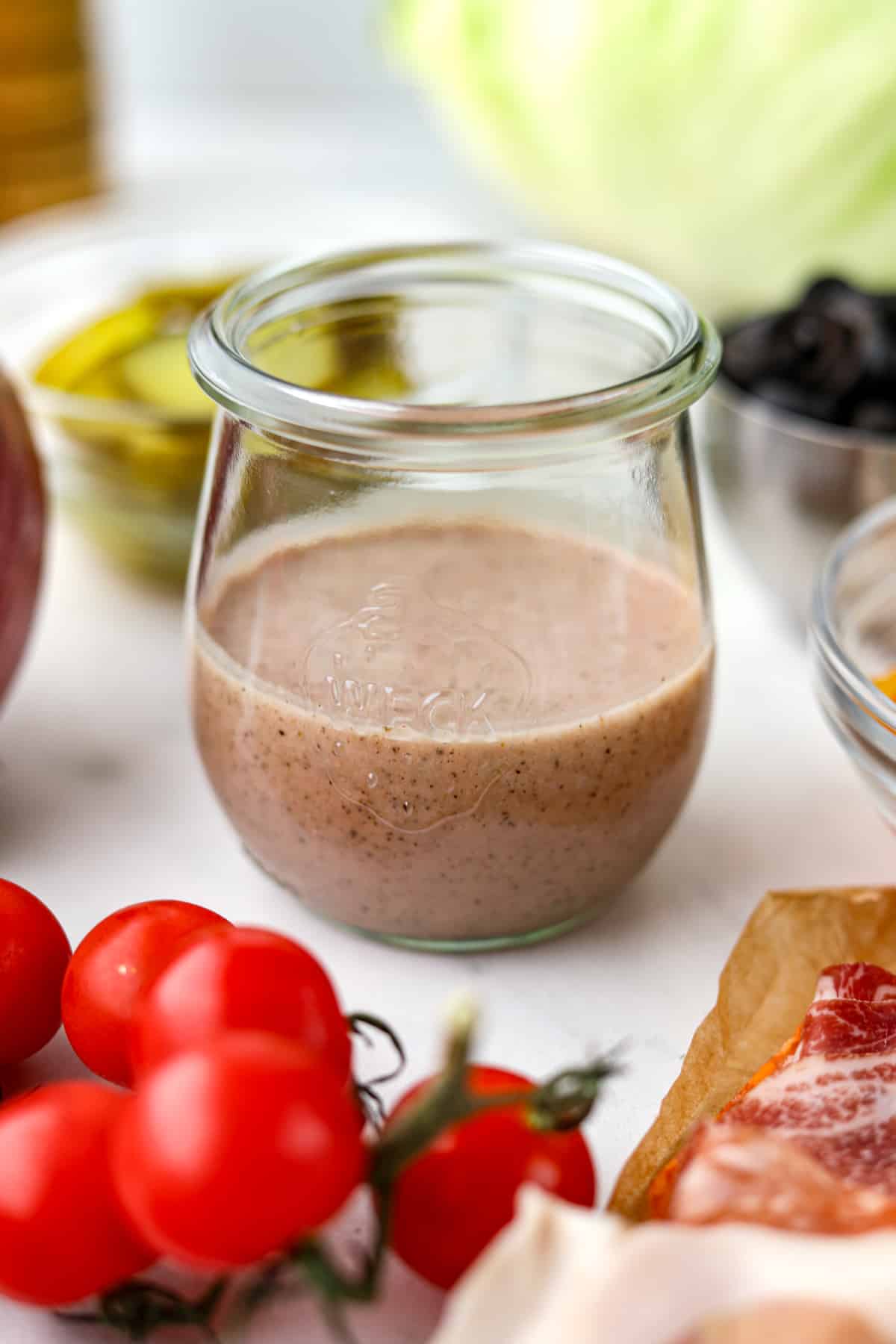 A small jar filled with Italian sub dressing, surrounded by cherry tomatoes, lettuce, etc.