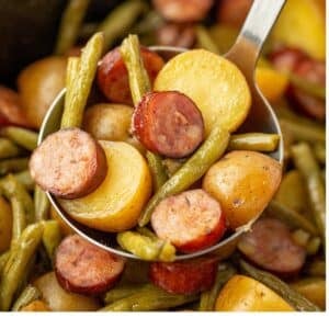 Scooping cooked potatoes, green beans, and smoked sausage from a slow cooker.