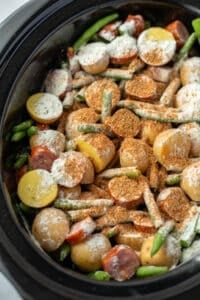A slow cooker filled with potatoes, green beans, and sausages sprinkled with seasoning.