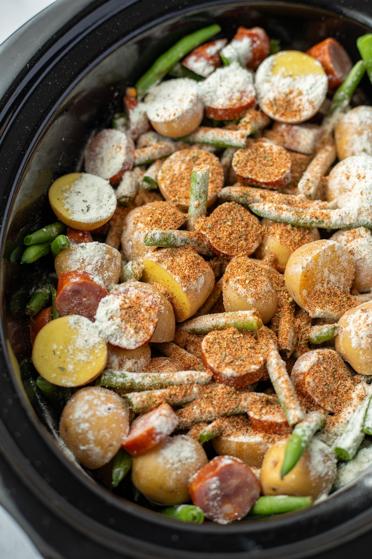 A slow cooker filled with potatoes, green beans, and sausages sprinkled with seasoning.