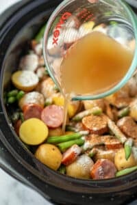 Pouring broth into a slow cooker filled with potatoes, green beans, and smoked sausage.
