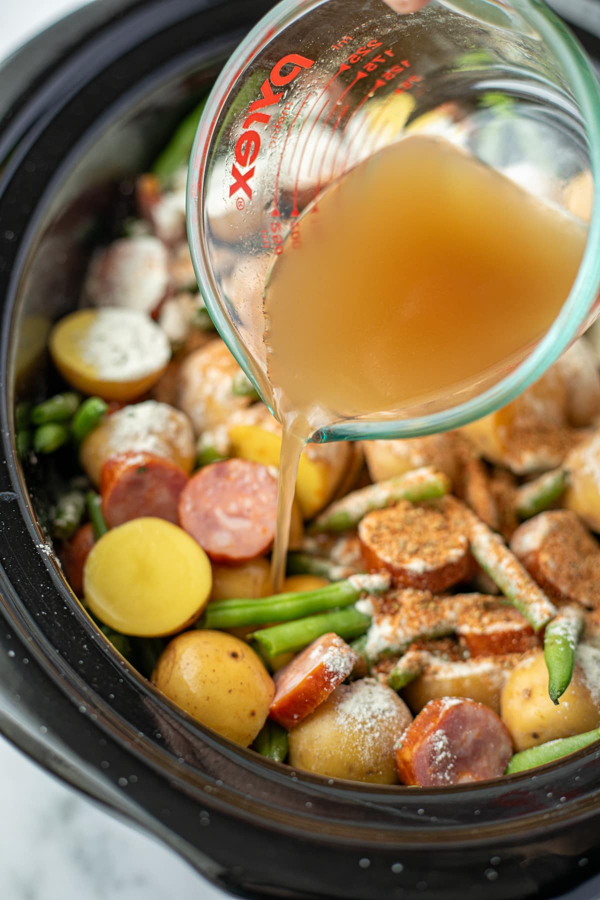 Pouring broth into a slow cooker filled with potatoes, green beans, and smoked sausage.
