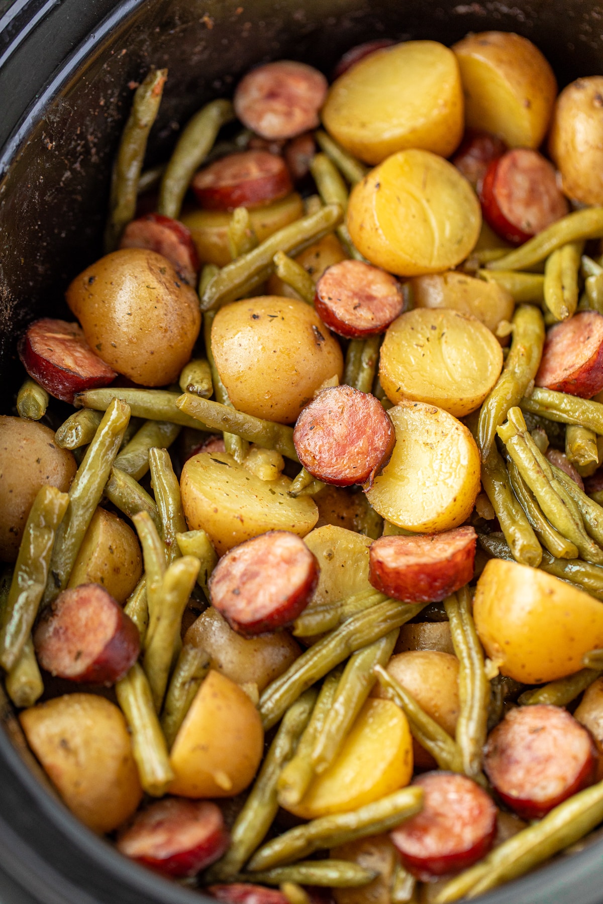 A slow cooker filled with cooked potatoes, green beans, and smoked sausage.